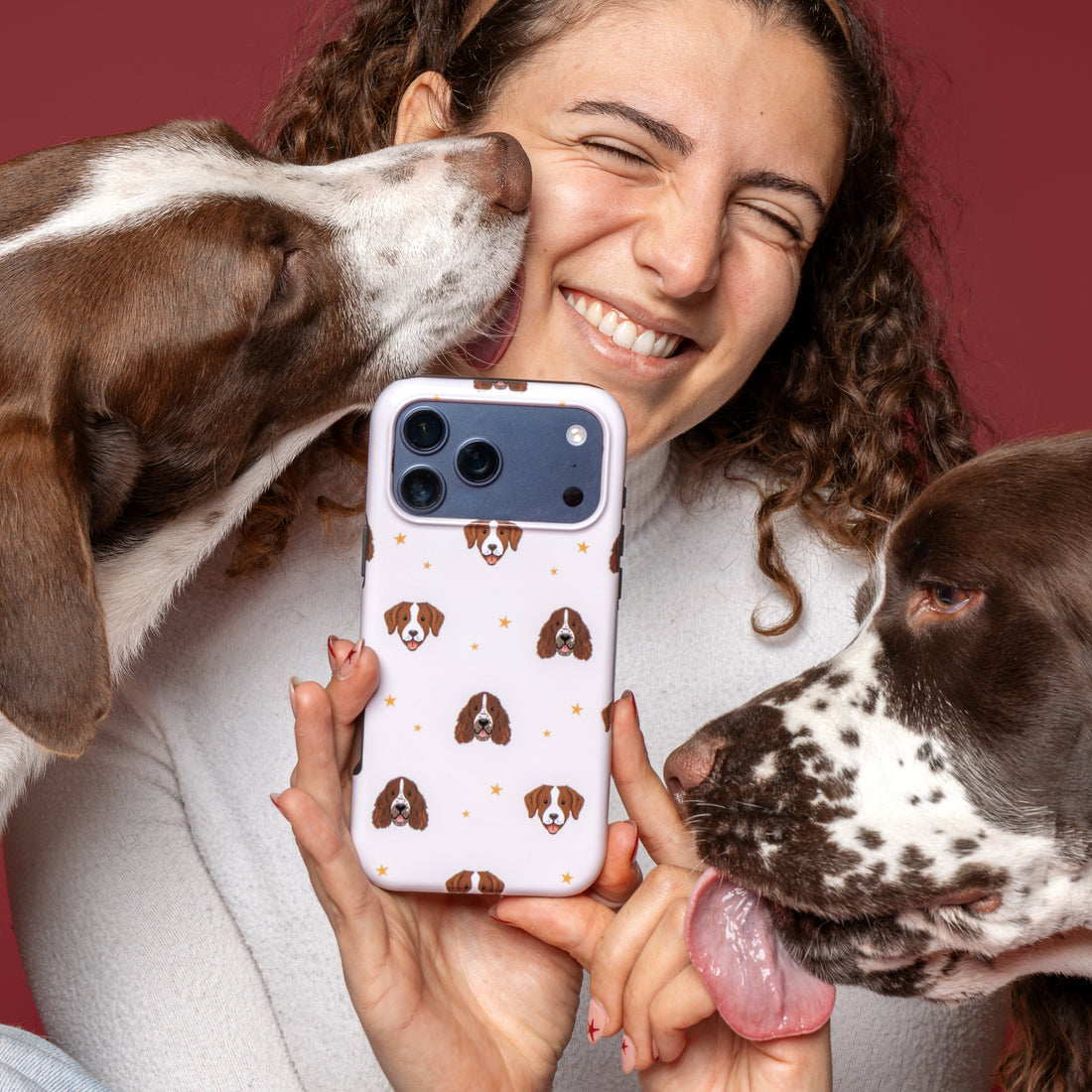 A woman holding a white phone case with a brown dog print pattern, surrounded by two brown and white dogs against a solid dark red background.