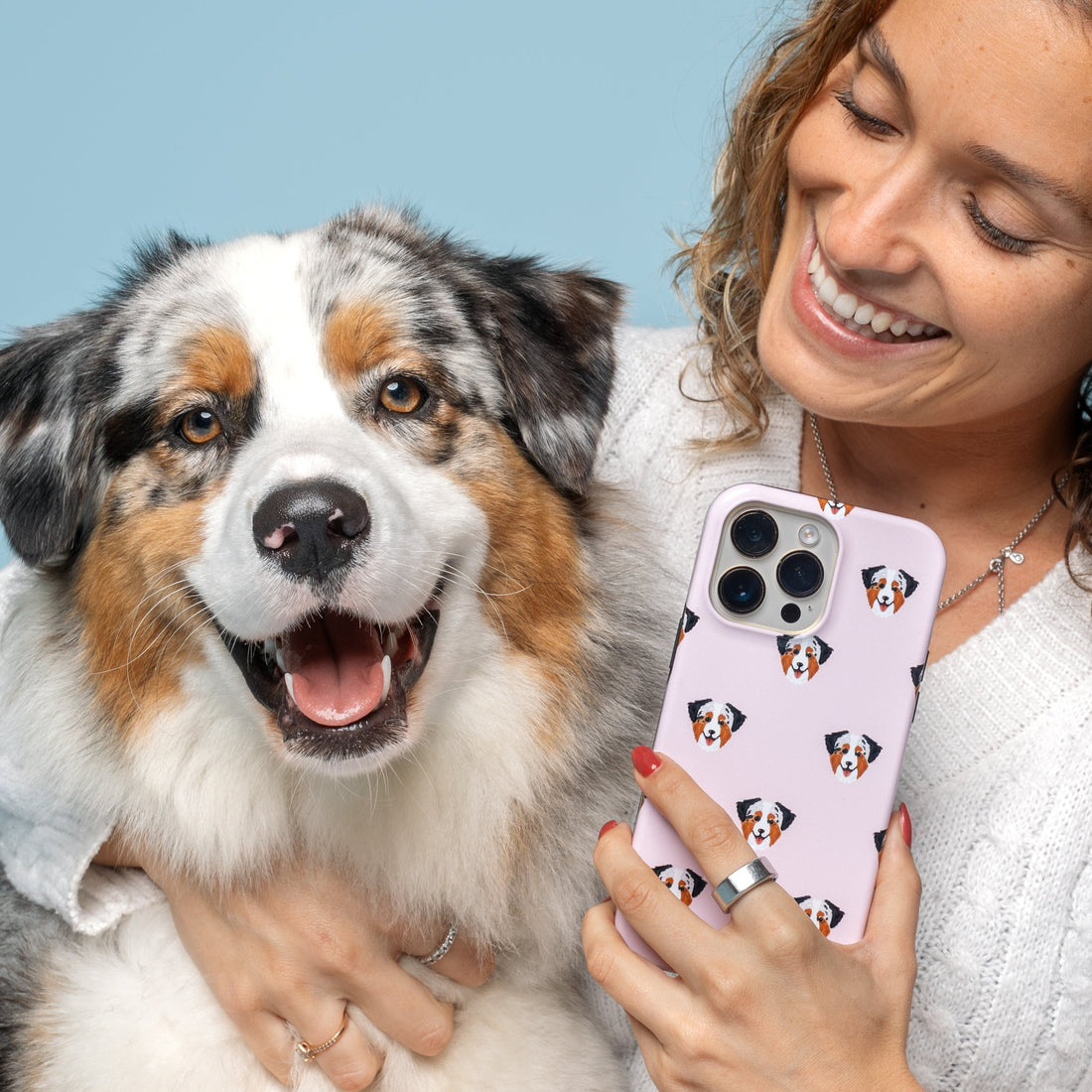 A woman in a white cable-knit sweater and blush pink pants holds a light pink phone case with a repeating dog face pattern, smiling as she looks at the screen. An Australian Shepherd dog sits beside her against a solid light blue background.